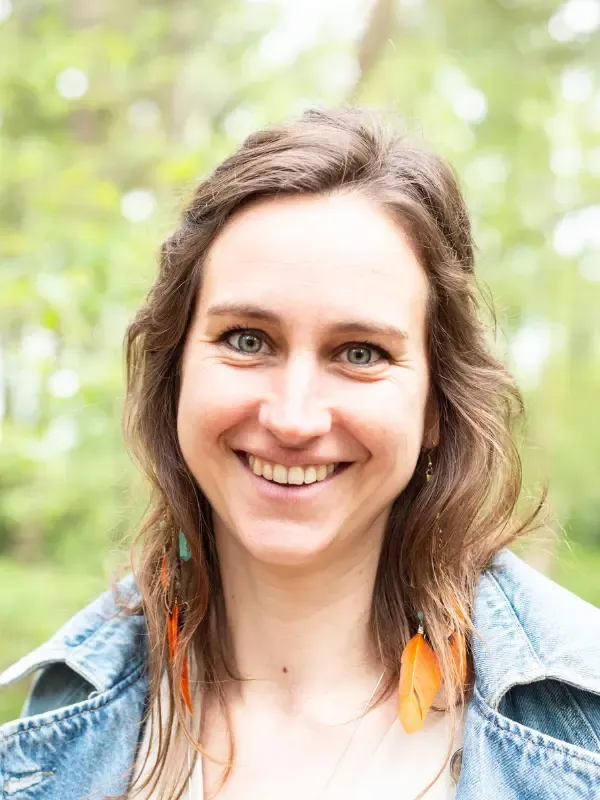 Closeup van Griet Verhaert in jeansjas, met sprekende oorbellen, tegen een bosrijke achtergrond